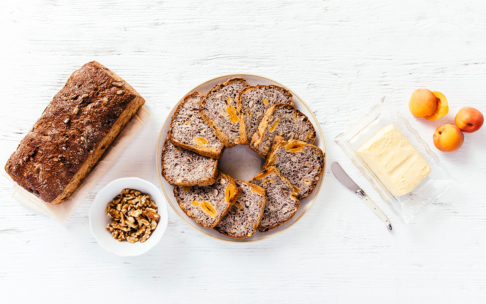 Ein Laib Nuss- und Früchtebrot, ein Teller mit geschnittenem Brot und getrockneten Aprikosen, eine Schale mit Walnüssen, drei frische Aprikosen und eine gläserne Butterdose mit einem Messer, alles auf einer weißen Holzfläche.