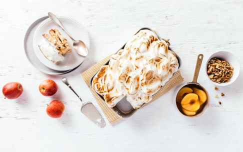 Ein rechteckiges gebackenes Dessert mit gebräuntem Baiser liegt auf einem weißen Tisch, umgeben von Äpfeln, einer Schale mit Walnüssen, einer Schale mit Dosenpfirsichen und einem in Scheiben geschnittenen Stück des Desserts auf einem Teller mit Gabel und Tortenheber.
