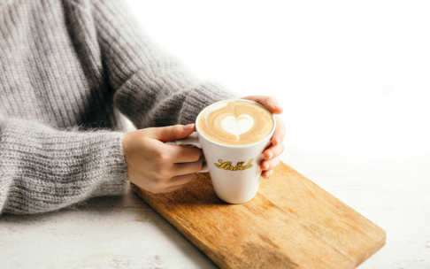 Eine Person in einem grauen Pullover hält eine weiße Tasse Milchkaffee mit herzförmiger Latte, die auf einem Holzbrett vor einem weißen Hintergrund ruht.