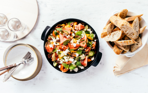 Ein frischer Salat mit Feta, Tomaten und Grünzeug in einer schwarzen Schale, daneben eine Schale mit Baguettescheiben, drei leere Gläser und gestapelte Teller mit Gabeln auf einer weißen Tischplatte.
