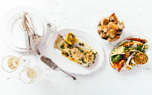 Ein weiß gedeckter Tisch mit einem Teller mit gekochtem Fisch, garniert mit Kräutern und Kapern, einer gegrillten Zitronenhälfte, einer Schüssel mit gemischtem geröstetem Gemüse, einer Schüssel mit Brotscheiben, zwei Gläsern Weißwein und Besteck auf gestapelten Tellern.