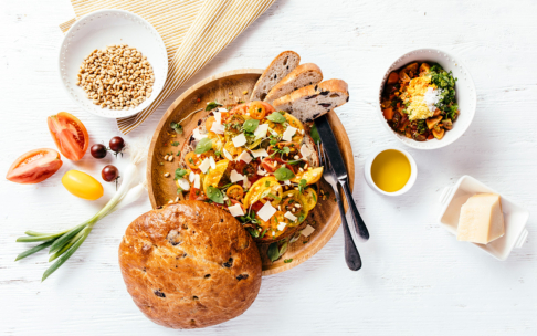 Ein Holztablett mit einem bunten Tomatensalat mit Käse und Kräutern, Brotscheiben, einem runden Laib, einer Schale mit Pinienkernen, geschnittenen Tomaten, grünen Zwiebeln, einer Schüssel mit Eintopf, Olivenöl und einem Käseblock auf einem weißen Tisch.