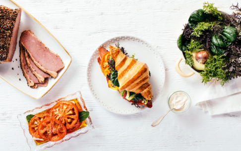 Ein Croissant-Sandwich mit Grünzeug und Schinken auf einem Teller, umgeben von Tomatenscheiben, Pastete, einer Schüssel mit gemischtem Grünzeug mit Avocado und einem kleinen Glas Soße auf einem weißen Tisch.