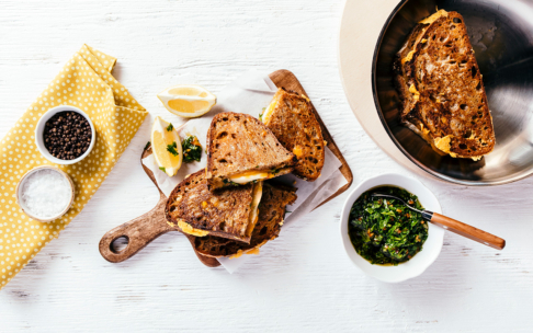 Gegrillte Käsesandwiches auf einem Holzbrett mit Zitronenspalten, einer Schale Chimichurri-Sauce, Salz, Pfeffer und einer gelben Serviette auf einem weißen Tisch. Ein weiteres Sandwich liegt in einer Metallpfanne.
