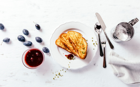 Ein weißer Teller mit zwei Scheiben Toast, garniert mit Marmelade und Kräutern, steht auf einem weißen Tisch. In der Nähe stehen ein Glas mit roter Marmelade, verstreute Pflaumen, ein Metallsieb, Besteck und eine strukturierte weiße Serviette.
