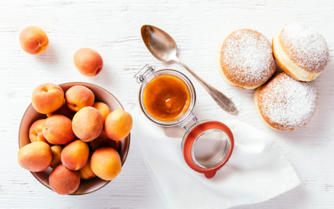 Eine Schale mit frischen Aprikosen, ein Glas Aprikosenmarmelade mit einem Löffel und drei mit Puderzucker bestreute Krapfen auf einer weißen Holzfläche.