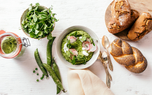 Eine Schale mit grüner Erbsensuppe, belegt mit Radieschenscheiben und Kräutern, umgeben von frischem Grün, ganzen Erbsen, einem Glas Pesto und verschiedenen Arten von knusprigem Brot auf einem weißen Holztisch. Zwei Löffel liegen daneben.