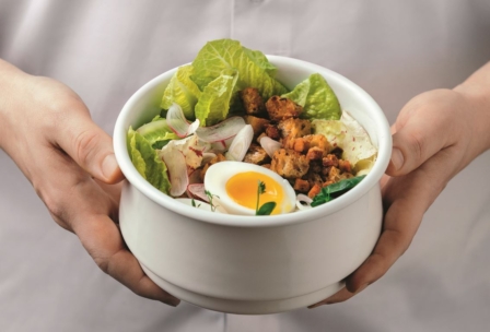 Eine Person mit einer weißen Schüssel, gefüllt mit Römersalat, Croutons, geschnittenen Radieschen, Zwiebeln und einem halbierten weichgekochten Ei.