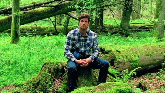Ein Mann in einem karierten Hemd und einer dunklen Hose sitzt auf einem moosbewachsenen, umgestürzten Baumstamm in einem üppigen, grünen Wald, der von Bäumen und Gestrüpp umgeben ist.