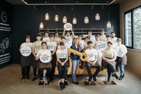 Eine Gruppe von Bäckereiangestellten, die Uniformen tragen und Schilder mit deutschem Text in der Hand halten, posieren gemeinsam in einem Innenraum. Eine Person in der Mitte hält einen großen, brezelförmigen Schlüssel in der Hand. Der Raum ist modern eingerichtet und hat große Fenster.