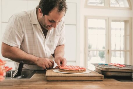 Ein Mann in einem kurzärmeligen Hemd verziert in einer hellen Küche eine Torte mit rosa und weiß gestreiftem Zuckerguss und arbeitet konzentriert an einem Holztisch.