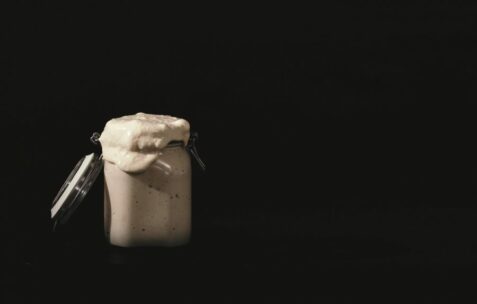 Ein mit einer cremigen Substanz gefülltes Glasgefäß läuft über und hat seinen Deckel geöffnet, vor einem dunklen, minimalistischen Hintergrund.