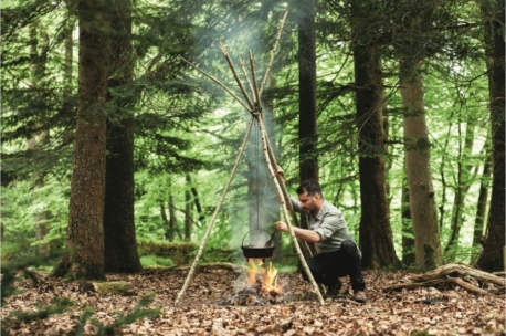 Eine Person hockt in einem grünen Wald und kümmert sich um einen schwarzen Topf, der auf einem Dreibein aus Stöcken über einem Lagerfeuer hängt, wobei Rauch in die Bäume aufsteigt.