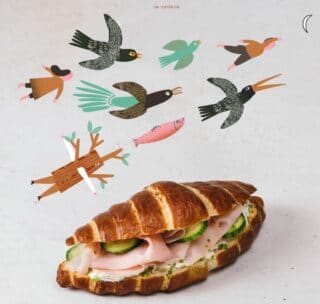 Ein Schinken-Gurken-Sandwich auf einem Croissant wird auf eine helle Fläche gelegt. Darüber fliegen bunte, illustrierte Vögel, ein pinkfarbener Fisch und ein Cartoon-Baum in die Höhe und sorgen für einen skurrilen und verspielten Effekt.