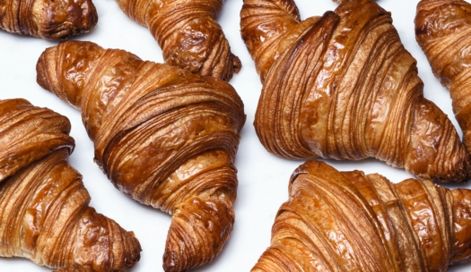 Goldbraune, blättrige Croissants sind auf einer weißen Oberfläche angeordnet, die ihre knusprige, geschichtete Textur und glänzende, gebackene Oberfläche zur Geltung bringt.