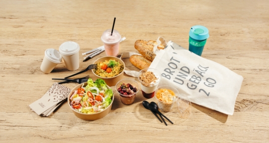Verschiedene Speisen zum Mitnehmen, darunter Salate, Joghurt-Parfaits, Getränke und Baguettes, stehen auf einem Holztisch mit einer Tragetasche mit der Aufschrift Brot und Gebäck 2 Go und mehreren Einwegbechern und -utensilien.