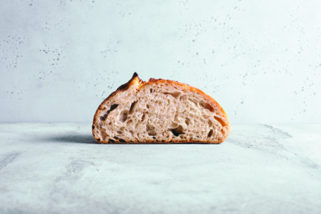 Eine einzelne Scheibe rustikales Sauerteigbrot mit einer knusprigen, goldenen Kruste und einer luftigen, offenen Krume liegt auf einer leicht strukturierten Oberfläche vor einem hellen Hintergrund.