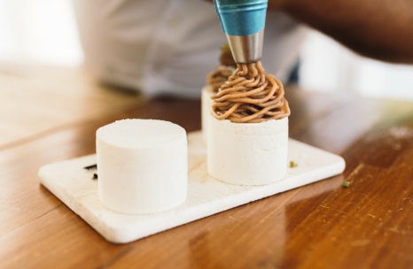 Eine Person schöpft braune Sahne auf ein zylindrisches weißes Dessert auf einem Schneidebrett, neben dem ein weiteres, nicht dekoriertes Dessert auf einem Holztisch steht.