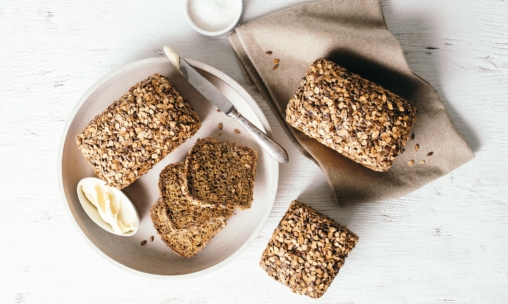 Drei Laibe Vollkornbrot, einige davon in Scheiben geschnitten, auf einem Teller mit einem Buttermesser und einer Schale mit Butter, alles auf einer weißen Holzfläche mit einer beigen Serviette.