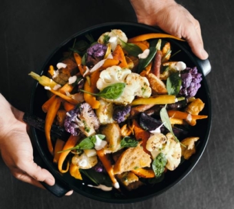 Eine Person hält eine schwarze Schüssel mit einem farbenfrohen Salat, der gerösteten Blumenkohl, Karotten, lila Gemüse, Blattgemüse, Croutons und einen Spritzer cremiges Dressing enthält.