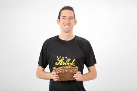 Ein lächelnder Mann in einem schwarzen Team Ströck-T-Shirt hält mit beiden Händen einen Laib Körnerbrot vor einem schlichten weißen Hintergrund.