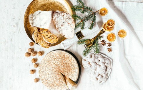 Ein festlich gedeckter Tisch mit Stollenbrot, Walnüssen, einem bestäubten Kuchen mit einer angeschnittenen Scheibe, Weihnachtsbaumzweigen, Goldschmuck, getrockneten Orangenscheiben und Sternanis auf einer weißen Holzfläche.