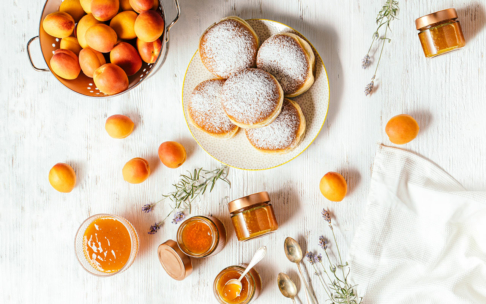Ein Teller mit Puderzucker-Donuts, frische Aprikosen, Gläser mit Aprikosenmarmelade und eine Schale mit Marmelade stehen auf einem weißen Tisch, auf dem Aprikosen und getrocknete Lavendelzweige verstreut sind.