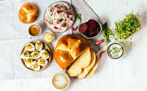 Ein gedeckter Tisch mit geschnittenem süßem Brot, hart gekochten Eiern mit Kräutern, Schinken, geschnittener Roter Bete, Senf, Mikrogrün, zwei Getränken und einer Tasse Kaffee auf einer weißen Holzfläche mit gestreiftem Tuch.