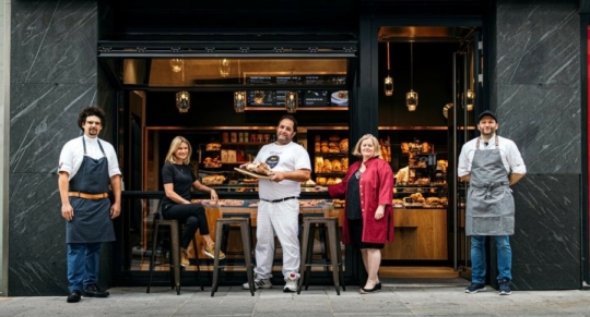 Fünf Personen stehen vor einer modernen Bäckerei; zwei tragen Schürzen, eine hält ein Brot in der Hand, eine trägt einen roten Mantel, und eine sitzt auf einem Hocker. Hinter ihnen ist das Innere der Bäckerei mit Backwaren zu sehen.