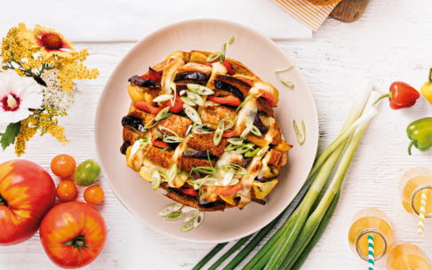 Ein Teller mit gebratenem Gemüse, überbacken mit geschmolzenem Käse und geschnittenen grünen Zwiebeln, umgeben von frischen Tomaten, Paprika, grünen Zwiebeln und einem Blumenstrauß auf einem weißen Holztisch.