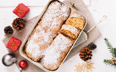 Zwei mit Puderzucker bestäubte traditionelle Stollen in einem Backblech, ein Laib teilweise aufgeschnitten. Das Blech ist umgeben von Tannenzapfen, Weihnachtsschmuck, Geschenkschachteln und kleinen goldenen Sternen.
