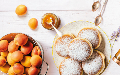 Auf einem weißen Holztisch steht ein Teller mit Donuts, die mit Puderzucker bestreut sind, neben einem Glas Aprikosenmarmelade mit einem Löffel, frischen Aprikosen in einem Sieb und zwei Löffeln.
