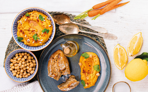 Ein Teller mit Hummus, Fladenbrot, Kichererbsen und Olivenöl, umgeben von Karotten, Zitronenscheiben und zwei dekorativen Hummus-Schalen, alles auf einem gewebten Tischset und einem weißen Holztisch.