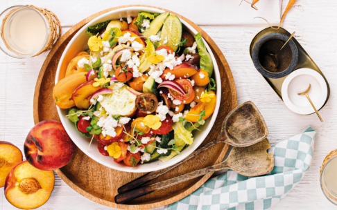 Ein bunter Salat mit Tomaten, Pfirsichen, Gurken, roten Zwiebeln, Schafskäse und essbaren gelben Blumen in einer weißen Schüssel auf einem Holztablett, daneben Salatbesteck aus Holz und Saftgläser.