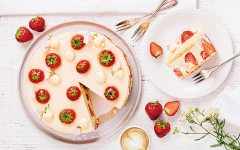 Ein runder Erdbeerkuchen mit Sahnetupfen und ganzen Erdbeeren oben drauf, ein Stück abgeschnitten und auf einem weißen Teller serviert, umgeben von frischen Erdbeeren, Gabeln, einer Tasse Kaffee und weißen Blumen auf einem weißen Tisch.