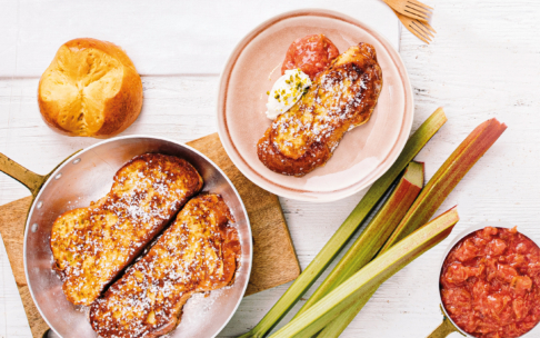 Mit Puderzucker bestäubter French Toast in einer Pfanne und auf einem Teller mit Schlagsahne und Obstkompott. In der Nähe stehen ein Brioche-Brötchen, frische Rhabarberstangen und ein Topf mit geschmortem Rhabarber auf einem weißen Holztisch.