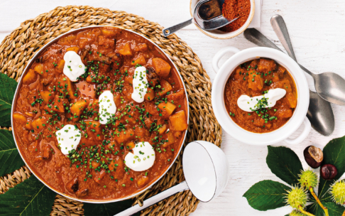 Auf einer geflochtenen Matte stehen eine Schüssel und ein Topf mit einem deftigen Eintopf mit Kartoffeln, der mit einem Klecks saurer Sahne und gehacktem Schnittlauch garniert ist. Um sie herum sind Löffel, eine Schale mit Gewürzen und grüne Blätter angeordnet.