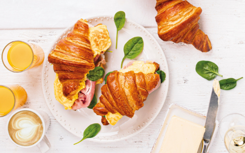 Ein Teller mit Croissant-Sandwiches, gefüllt mit Rührei, Schinken und Spinat, umgeben von weiteren Croissants, Spinatblättern, zwei Gläsern Orangensaft, einer Tasse Kaffee mit Latte Art und einer Schale mit Butter und einem Messer auf einem weißen Tisch.