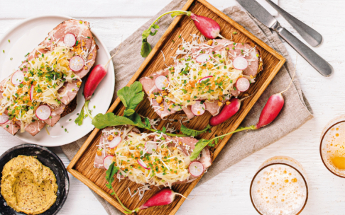 Ein Holztablett und ein weißer Teller mit Brotscheiben, belegt mit Fleisch, geriebenem Käse, geschnittenen Radieschen, Kräutern und serviert mit ganzen Radieschen. In der Nähe stehen Senf in einer Schüssel und Biergläser auf einem weißen Tisch.