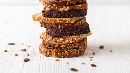 Ein Stapel gemischter Mehrkornbrotscheiben liegt auf einer weißen Holzunterlage, auf der die Samen verstreut sind. Das Brot weist sichtbare Samen wie Sesam und Sonnenblumen auf der Kruste auf.