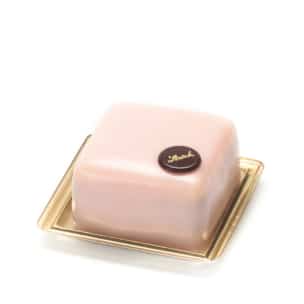 Eine kleine, quadratische rosa Mousse-Torte mit glatter, glänzender Oberfläche steht auf einem goldenen Tablett. Die Torte ist mit einer runden Schokoladenscheibe mit goldener Aufschrift gekrönt.