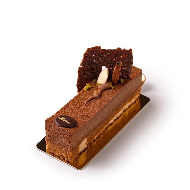 Eine rechteckige Schokoladenmousse-Torte mit geschichteter Füllung, verziert mit Schokoladenstückchen, einer Zimtstange und einem kleinen Sahnestrudel, auf einem Metalltablett vor weißem Hintergrund.