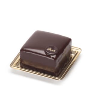 Eine quadratische Schokoladentorte mit glänzender Schokoladenglasur, gekrönt von einer kleinen runden Schokoladenscheibe mit dem Schriftzug Sacher, präsentiert auf einem goldenen Tablett.