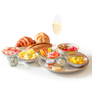 Ein Frühstücksbuffet mit Croissant, Brötchen, Aufschnitt, Käse, geschnittenem Gemüse, Rührei, Joghurt mit Müsli, kleinen Marmeladengläsern, Butter und einem Glas Sekt, alles auf weißem Geschirr.