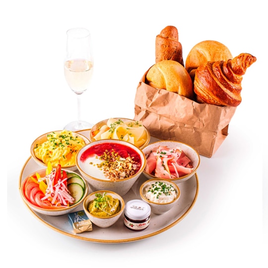 Ein Frühstücksaufstrich mit Rührei, Käse, Schinken, geschnittenem Gemüse, Joghurt mit Müsli und Beeren, Dips, Butter, Marmelade, Croissants, Brötchen in einer Papiertüte und einem Glas Sekt auf weißem Hintergrund.