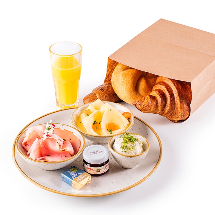 Ein Frühstücksbrot mit einem Teller mit Schinkenscheiben, Käse, Butter, einem Glas Marmelade und einer Schale mit cremigem Aufstrich, daneben ein Glas Orangensaft und eine Papiertüte mit einem Croissant und einem Brötchen.
