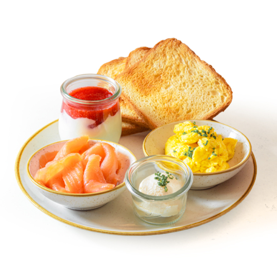 Ein Frühstücksteller mit zwei Scheiben Toast, Rührei mit Schnittlauch, Räucherlachs, einem kleinen Glas Joghurt mit Erdbeersauce und einer Schale Frischkäse.