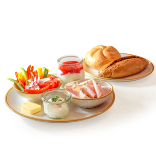 Ein Frühstücksaufstrich mit geschnittenem Schinken, buntem Gemüsesalat, Butter, einem Brötchen, einem braunen Baguette, Joghurt mit Obst und einem kleinen Glas Frischkäse, alles auf beigen Tellern vor weißem Hintergrund.