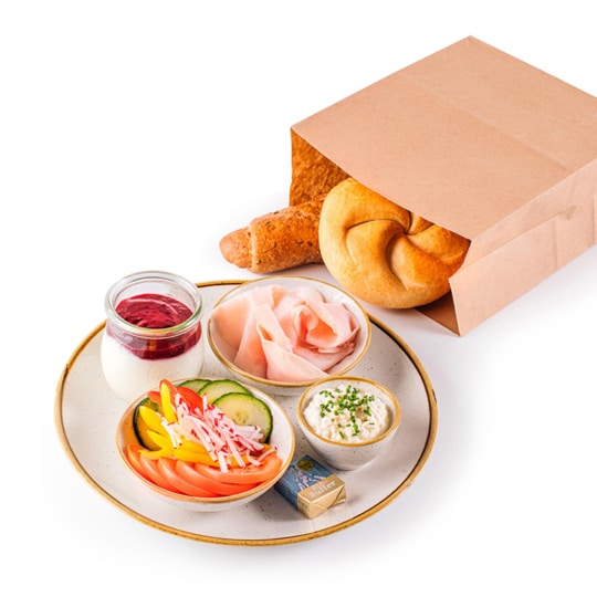 Ein Frühstücksset mit einer Papiertüte, Brötchen, einem Teller mit Schinken, geschnittenem Gemüse, Frischkäse, einem kleinen Glas Joghurt mit Fruchtsauce und einer Portion Butter auf weißem Hintergrund.