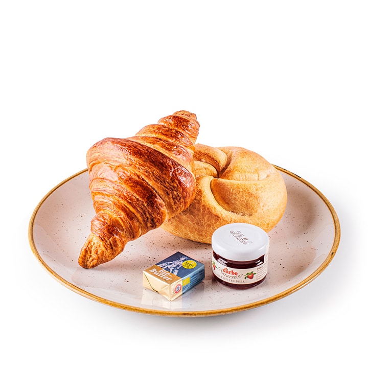Ein Teller mit einem Croissant, einem runden Brötchen, einem kleinen Päckchen Butter und einem Mini-Glas Erdbeermarmelade, alles fein säuberlich auf einem hellen Keramikteller vor weißem Hintergrund angeordnet.